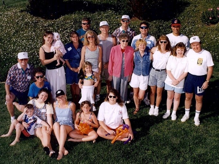 Group of people outdoors on grass, posing for a photo, some holding children and toys, in casual summer clothing.