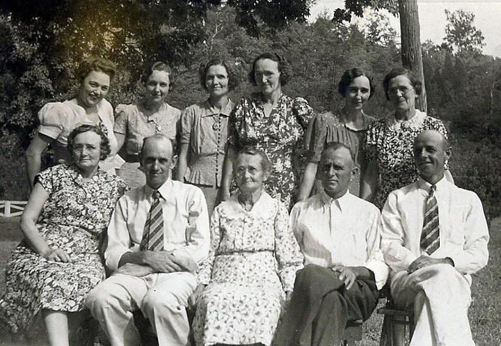 Black and white photo of ten people, five women and five men, outdoors with trees and a wooden pole in the background. The women are wearing dresses, and the men are wearing shirts and ties. The group is posing, some sitting and others standing.
