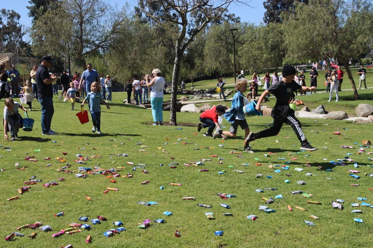 People participating in an Easter egg hunt in a park on a sunny day. Children are collecting colorful eggs scattered on the grass while adults watch and take photos.