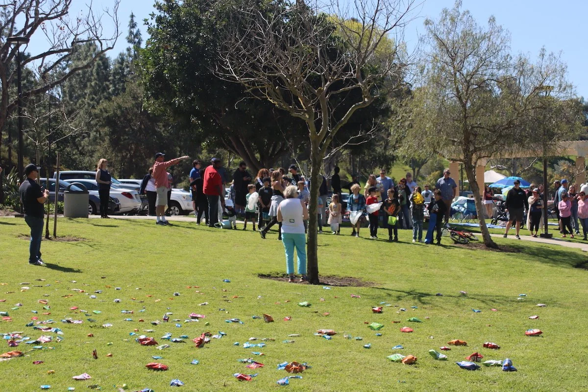 People gathered in a park, possibly during a celebration or event, with a tree and fallen candy wrappers on the grass.