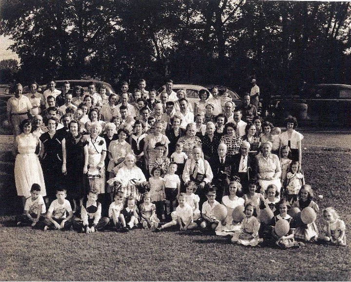 A large group of children and adults posing outdoors on a grassy field with trees and vintage cars in the background, in black and white.