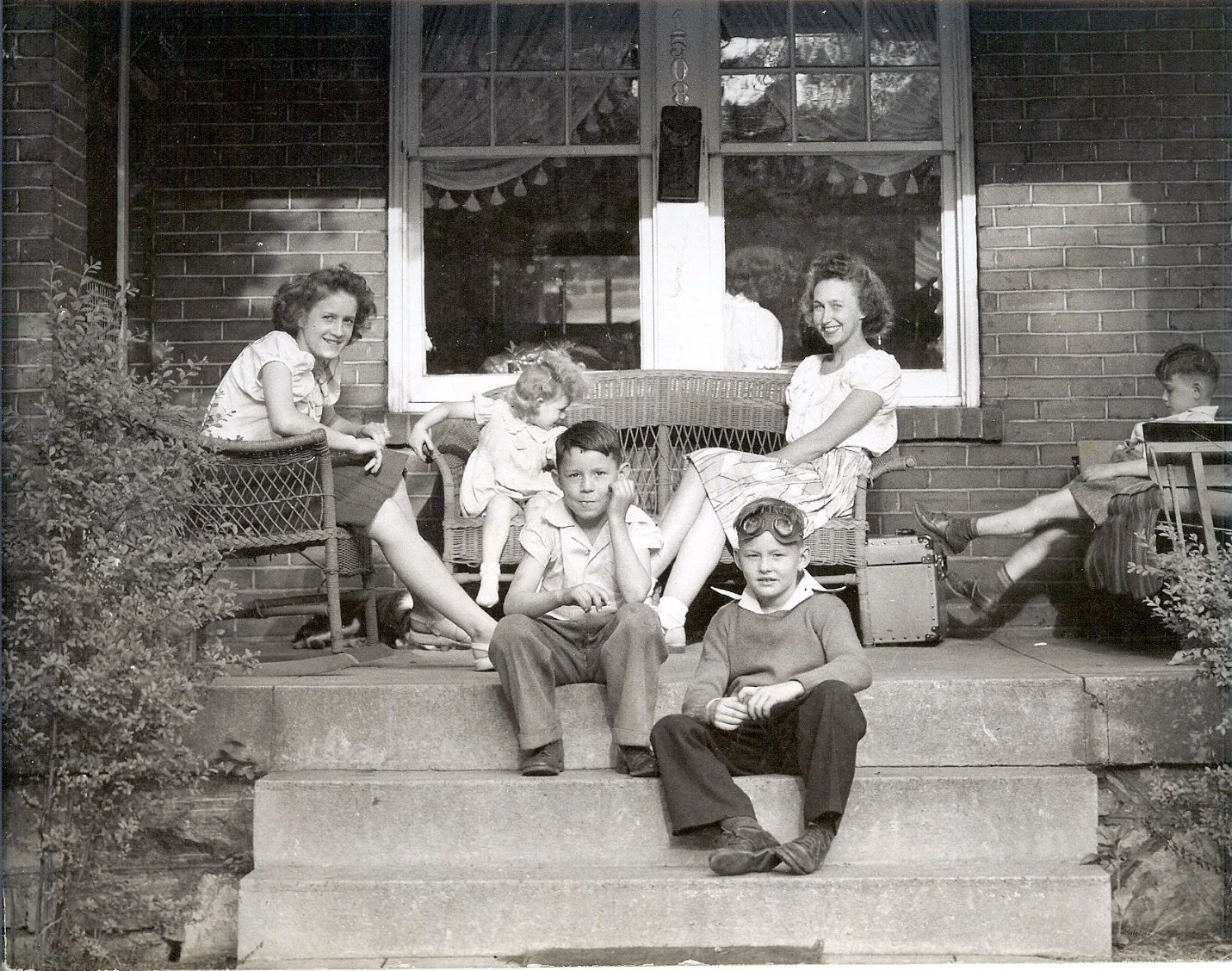 A black and white photo of six children sitting on the front steps of a brick house, with two women sitting on chairs behind them. The children appear to be posing for the photo, and some are wearing glasses or goggles. The scene looks to be from a p