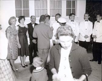 A group of men and women dressed in formal attire, gathered outdoors, with some people engaged in conversations.