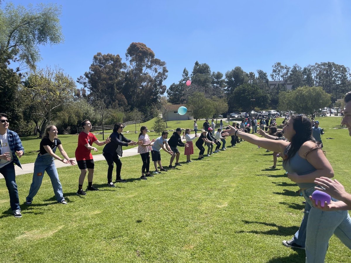 People participating in an outdoor game of tug-of-war on a grassy field, with some players holding colorful balloons and pulling on the rope while others cheer in the background.