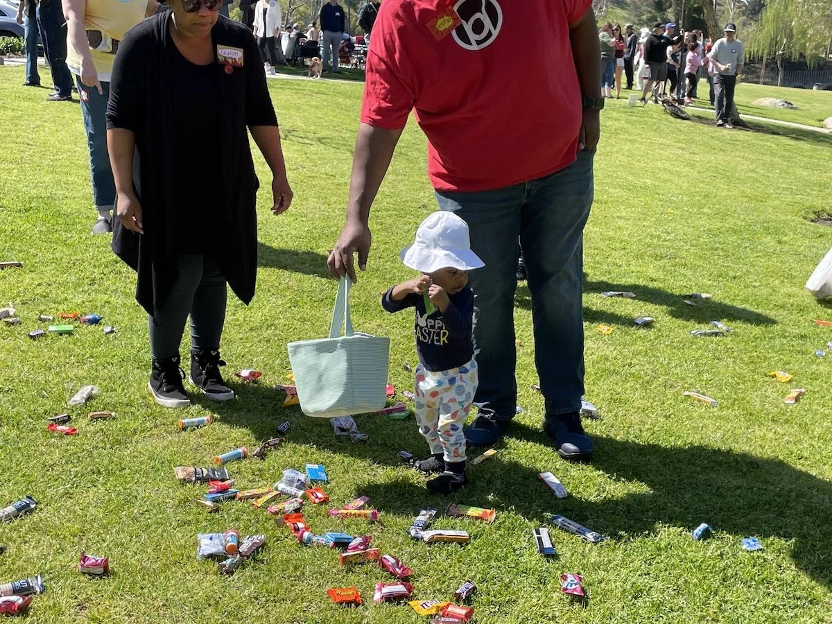 A young child wearing a white sun hat and colorful pants is walking on grass with scattered candy wrappers, while an adult in a red shirt holds the child's hand and a woman in black looks on. There are more people in the background on a sunny day.
