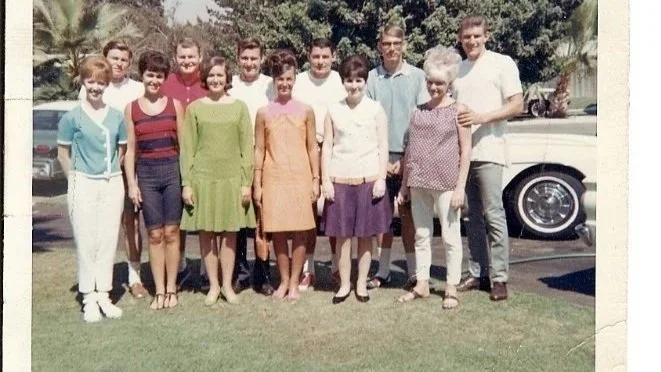A group of ten women and men standing outdoors on grass, posing for a photo with a vintage white car in the background, under a partly cloudy sky.