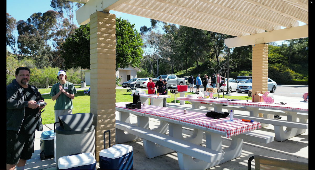 People gathering at a covered pavilion with picnic tables, some preparing food and others socializing, in a park with cars and trees in the background.