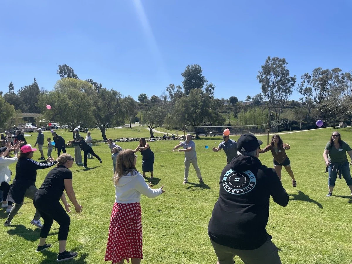 People playing a game of dodgeball on a grassy field on a sunny day with trees and a blue sky in the background.