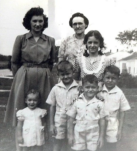 Black and white photo of two women and four children outdoors, with houses in the background.