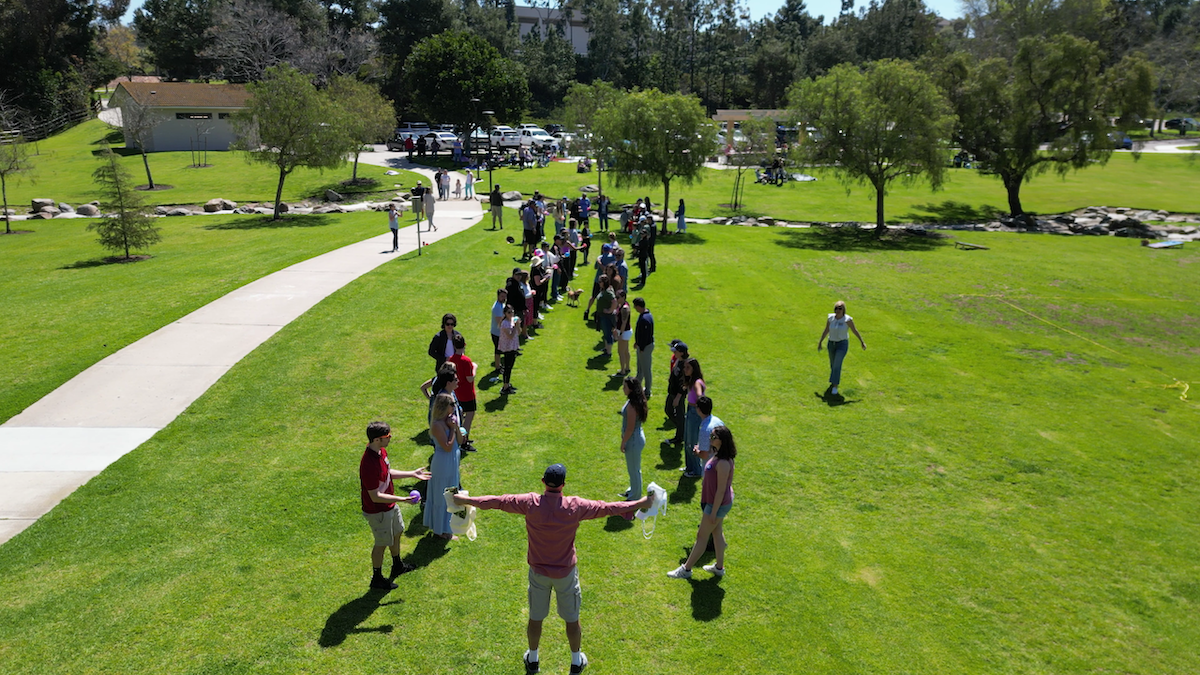 People outdoors standing in a line on a grassy area with trees, sidewalks, and a building in the background.
