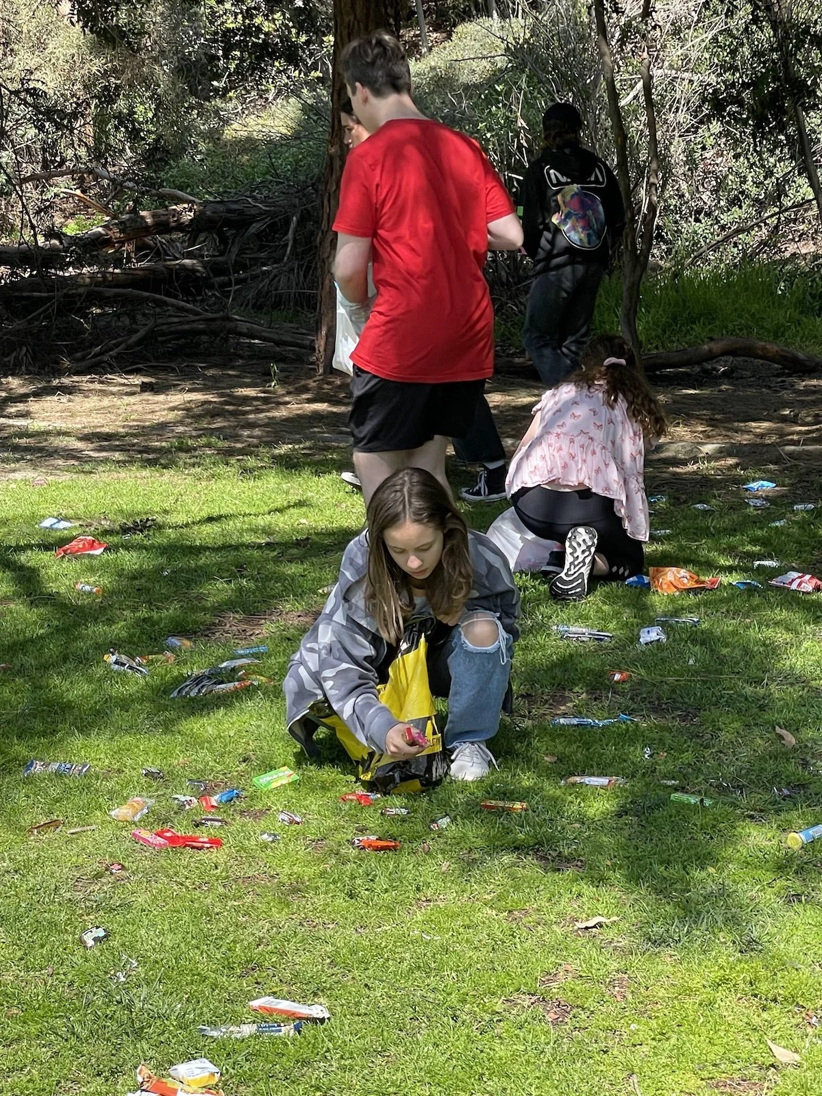 Children picking up trash in a park, with litter scattered on the grass, surrounded by trees.
