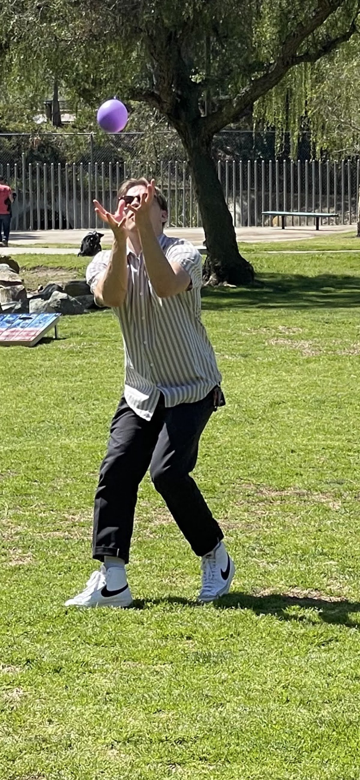 Person wearing a striped shirt and black pants playing with a purple balloon outdoors on a sunny day.