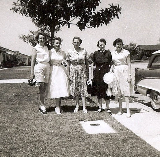 Five women standing outdoors on a lawn, dressed in vintage mid-20th-century clothing, with a tree and a car in the background.