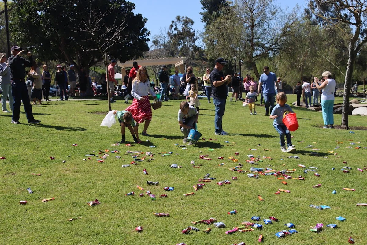 People celebrating Easter, children with buckets collecting candy on a grassy field, Easter eggs and candy scattered on the grass, trees, and a pavilion in the background.