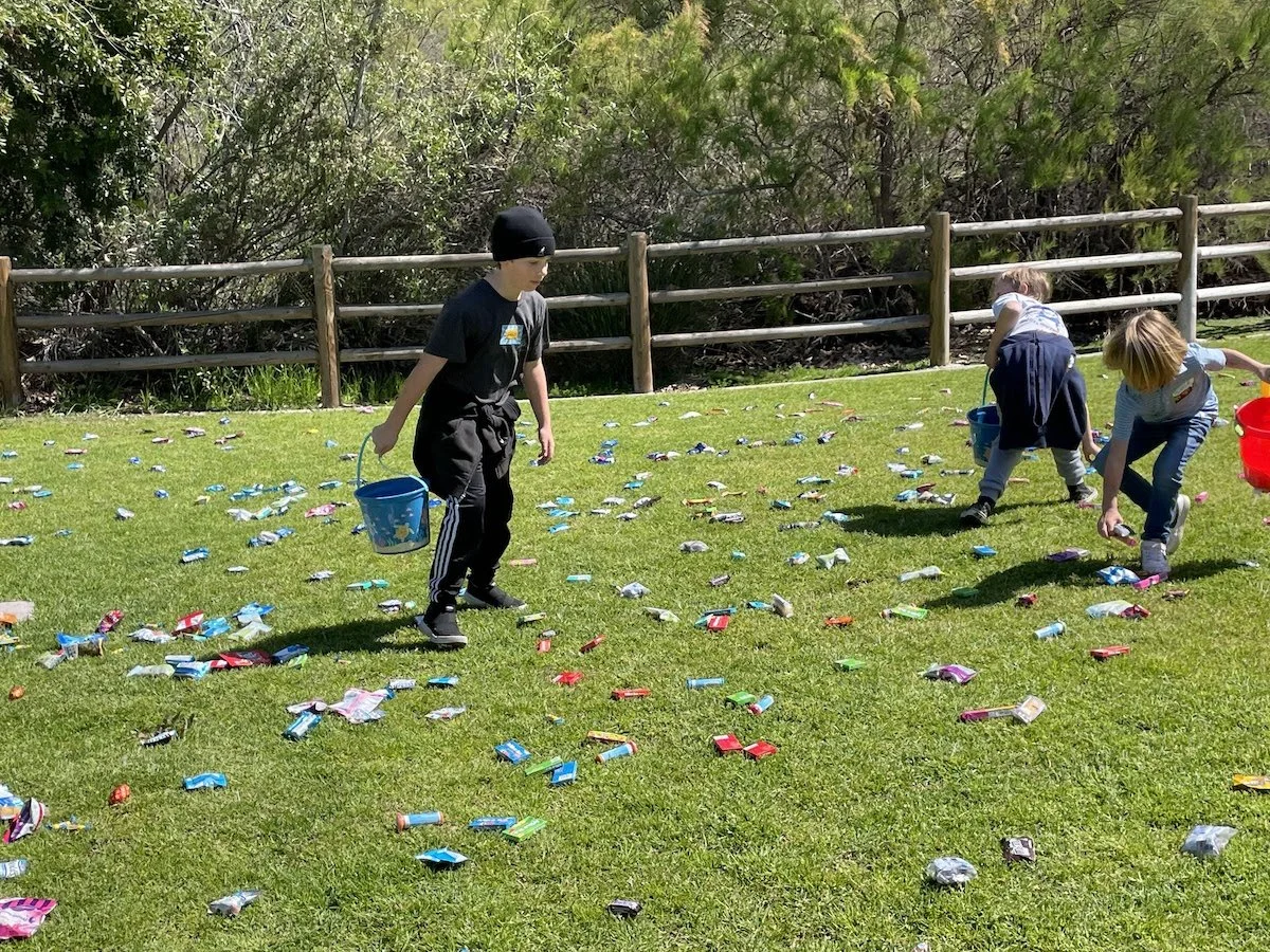 Children participating in an outdoor egg hunt, picking up colorful candy or plastic eggs on a grassy field, with a wooden fence and bushes in the background.