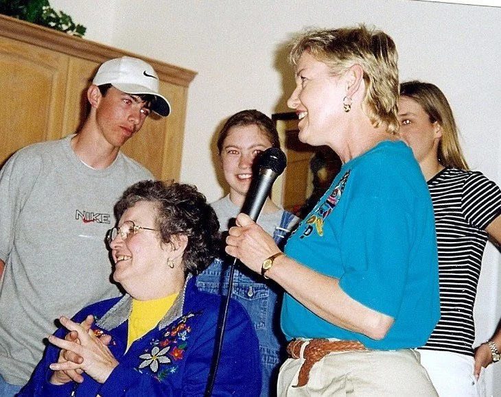A group of people gathered at an indoor event; a woman with a blonde haircut is speaking into a microphone, smiling and wearing a blue shirt and beige pants, while seated woman with glasses, a blue jacket, and a yellow shirt is smiling and clapping. 