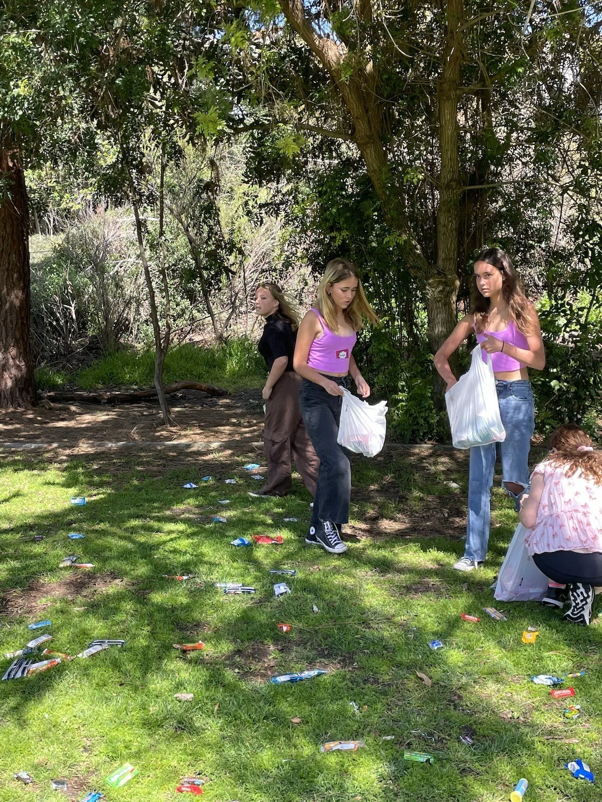 Four young women cleaning up litter in a wooded outdoor area, holding trash bags, with scattered trash on the grass.