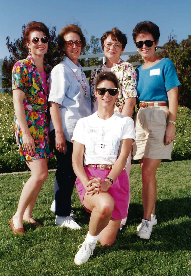 Five women outdoors on a sunny day, all wearing sunglasses, posing for a group photo on a grassy area with trees and a clear blue sky in the background.