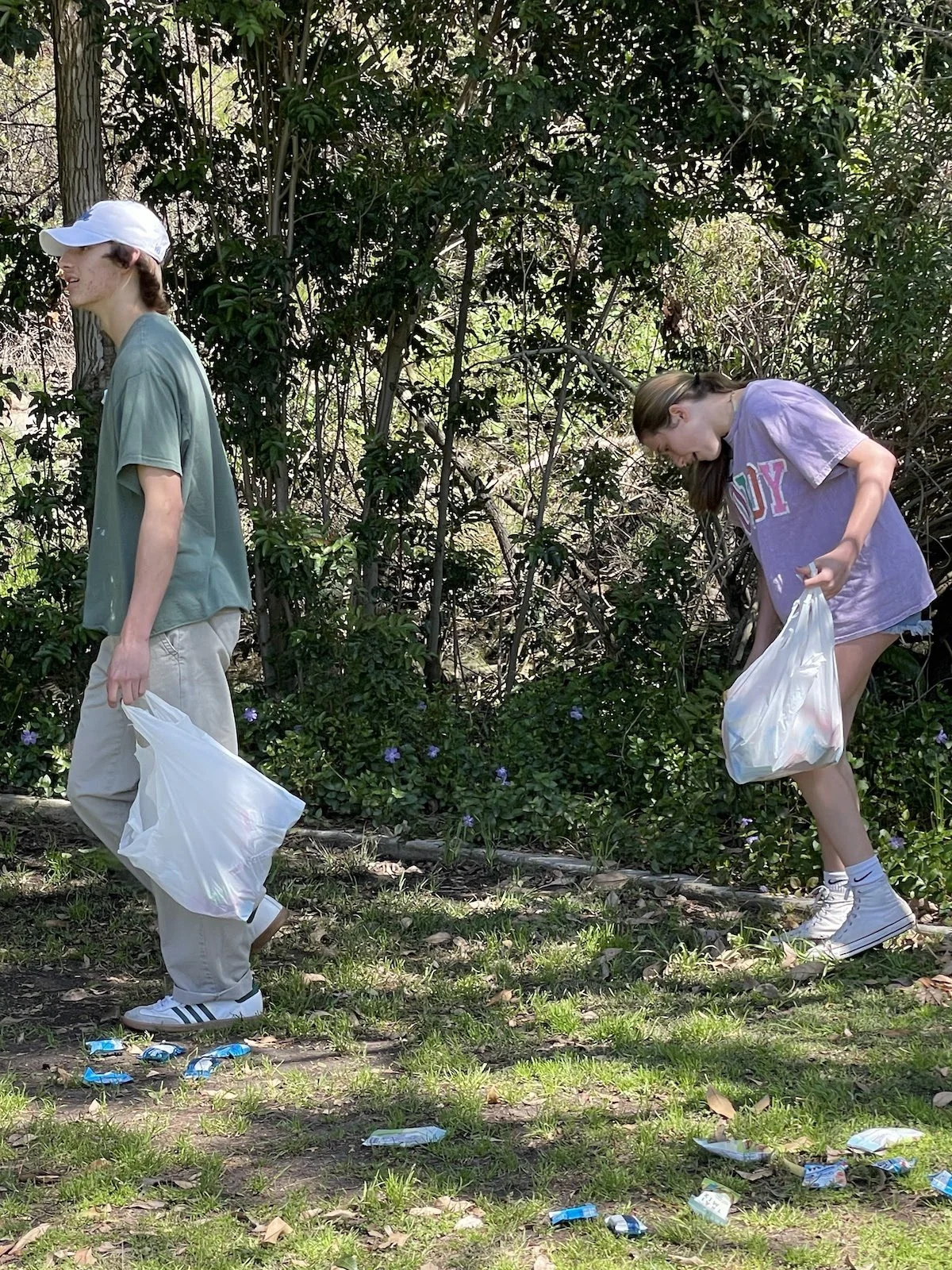 Two young women participating in a litter cleanup outdoor activity in a green area, one clipping litter with a trash bag in her hand, the other bent over collecting trash, trees and shrubs in background, scattered trash on ground.
