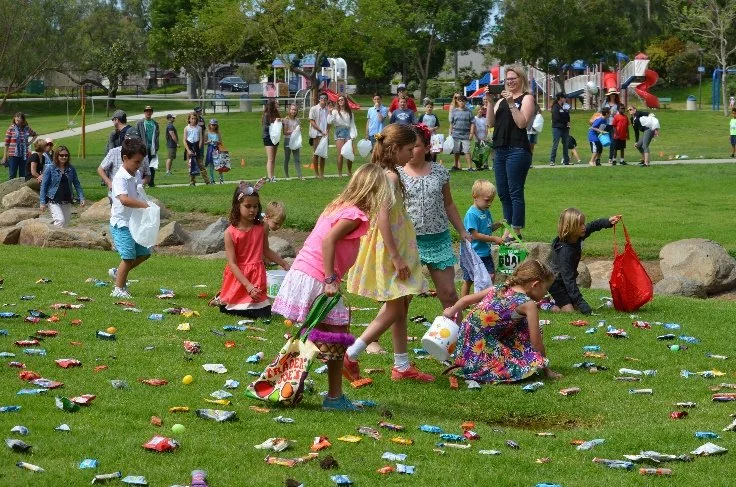 Children participating in an Easter egg hunt in a park, with many colorful eggs scattered on the grass and families watching in the background.