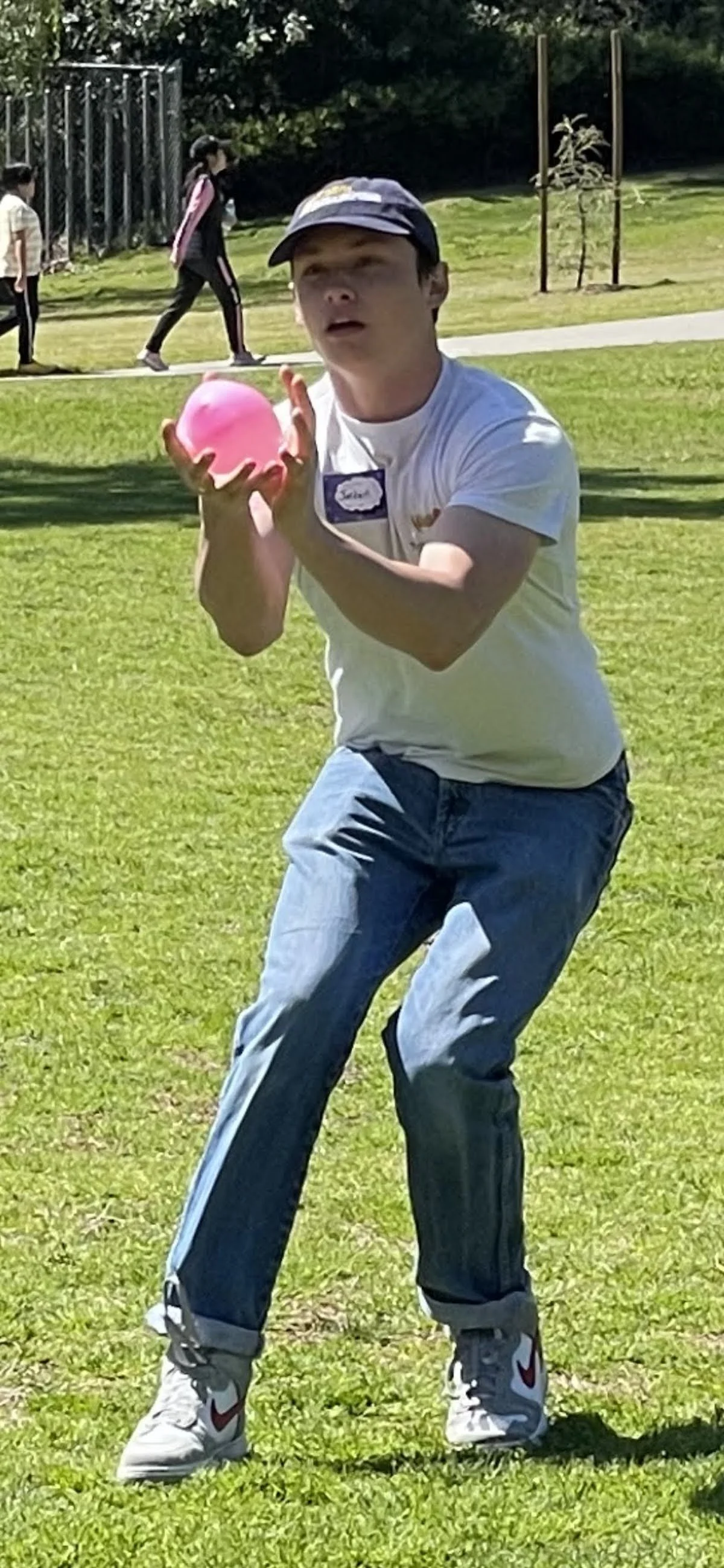 A young man wearing a white t-shirt, blue jeans, white Nike sneakers, and a cap is holding a pink ball with both hands, preparing to throw it during a game on a grassy field. In the background, two women are walking along a path, with a fence and tre