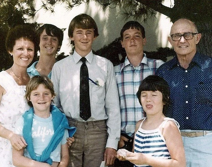 Group of seven people, including adults and children, smiling and posing outdoors under a tree.