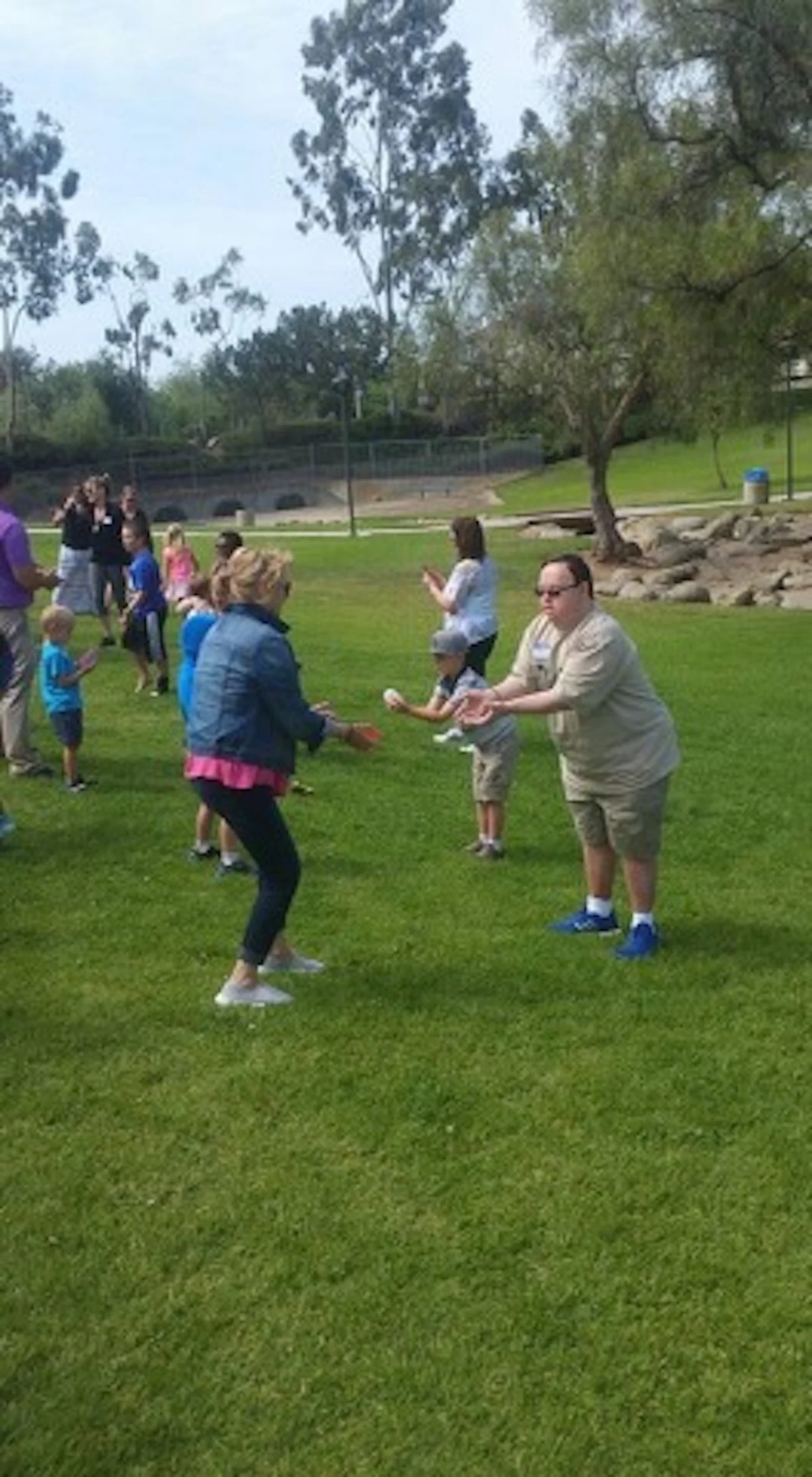 People outdoors in a park holding papers, engaging in a group activity or game on the grass, with trees and a fence in the background.