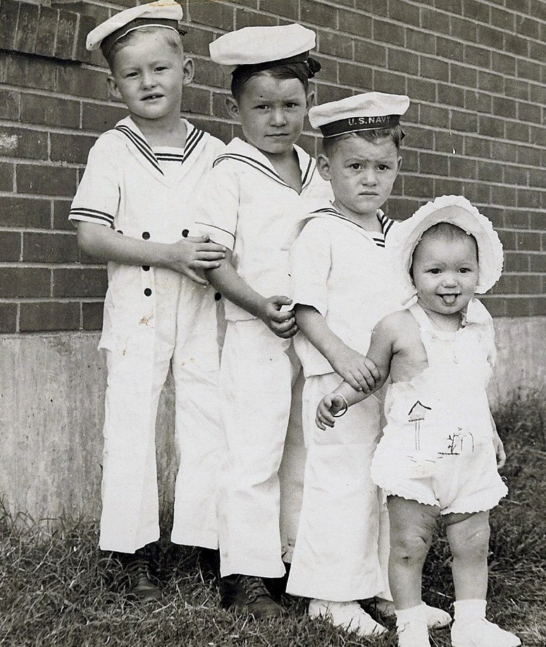 Four children dressed in sailor outfits standing outside against a brick wall, with the youngest girl wearing a sun hat.