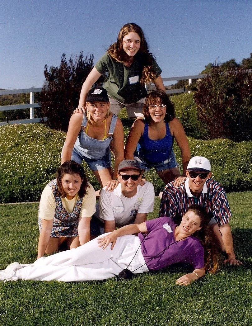 A group of six young friends posing outdoors on grass, with a woman lying down in front, two women and two men in the middle, and a woman in green behind, under a clear blue sky.