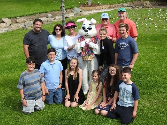 A group of children and adults posing outdoors on grass with a bunny mascot in the center wearing a colorful shirt and sunglasses.