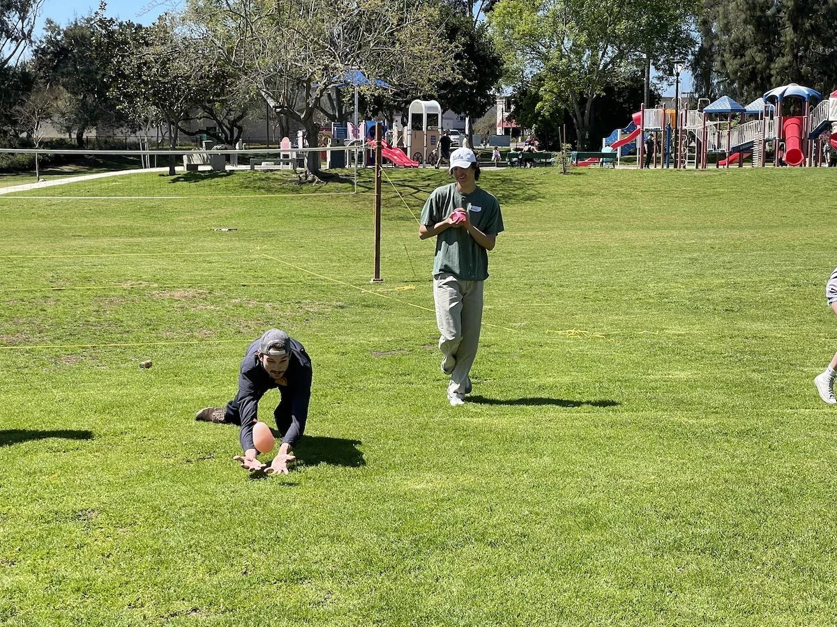 Child falling on grass during a game, with a woman standing nearby, at a park with play structures in the background.