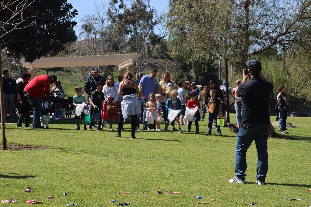 Group of children and adults participating in an outdoor egg hunt event on a grassy area with trees, with a man taking a picture and colorful eggs on the ground.