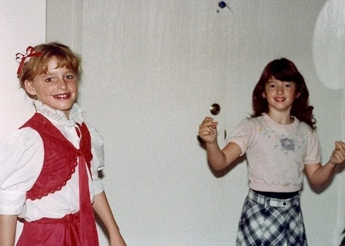 Two young girls standing indoors, smiling, with one girl holding a small object in her hand.