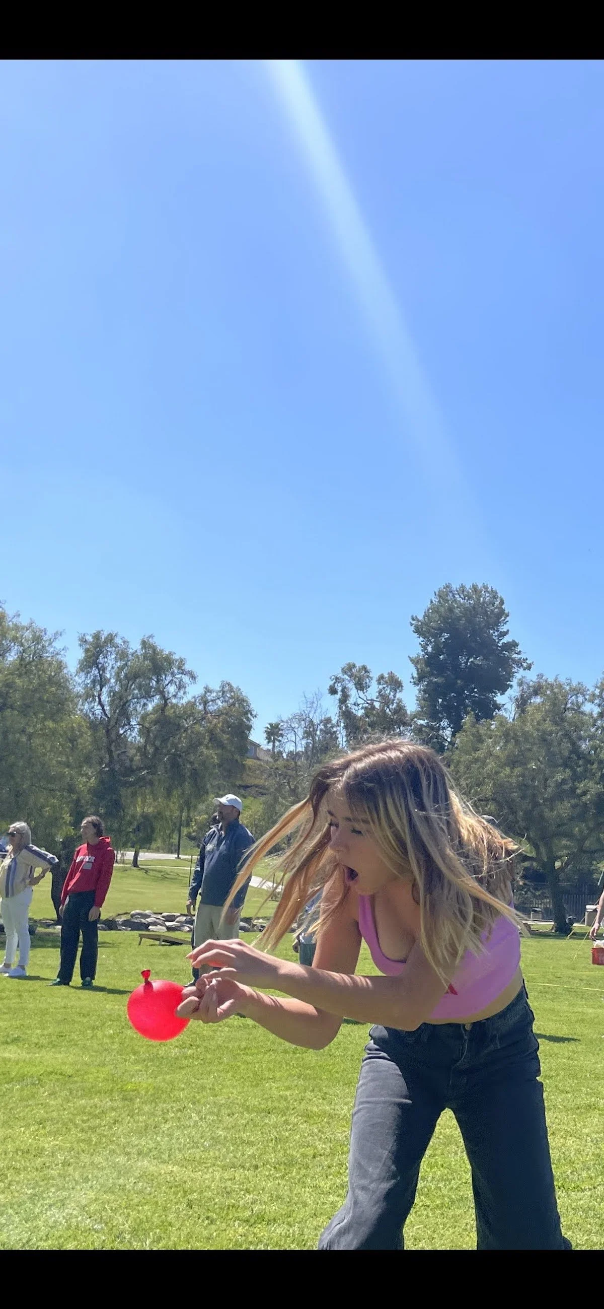 A young woman with long blonde hair, wearing a pink tank top and blue jeans, is outdoors on a sunny day, participating in a balloon-popping game by sitting and holding a red balloon between her knees. Several other people are in the background on a g