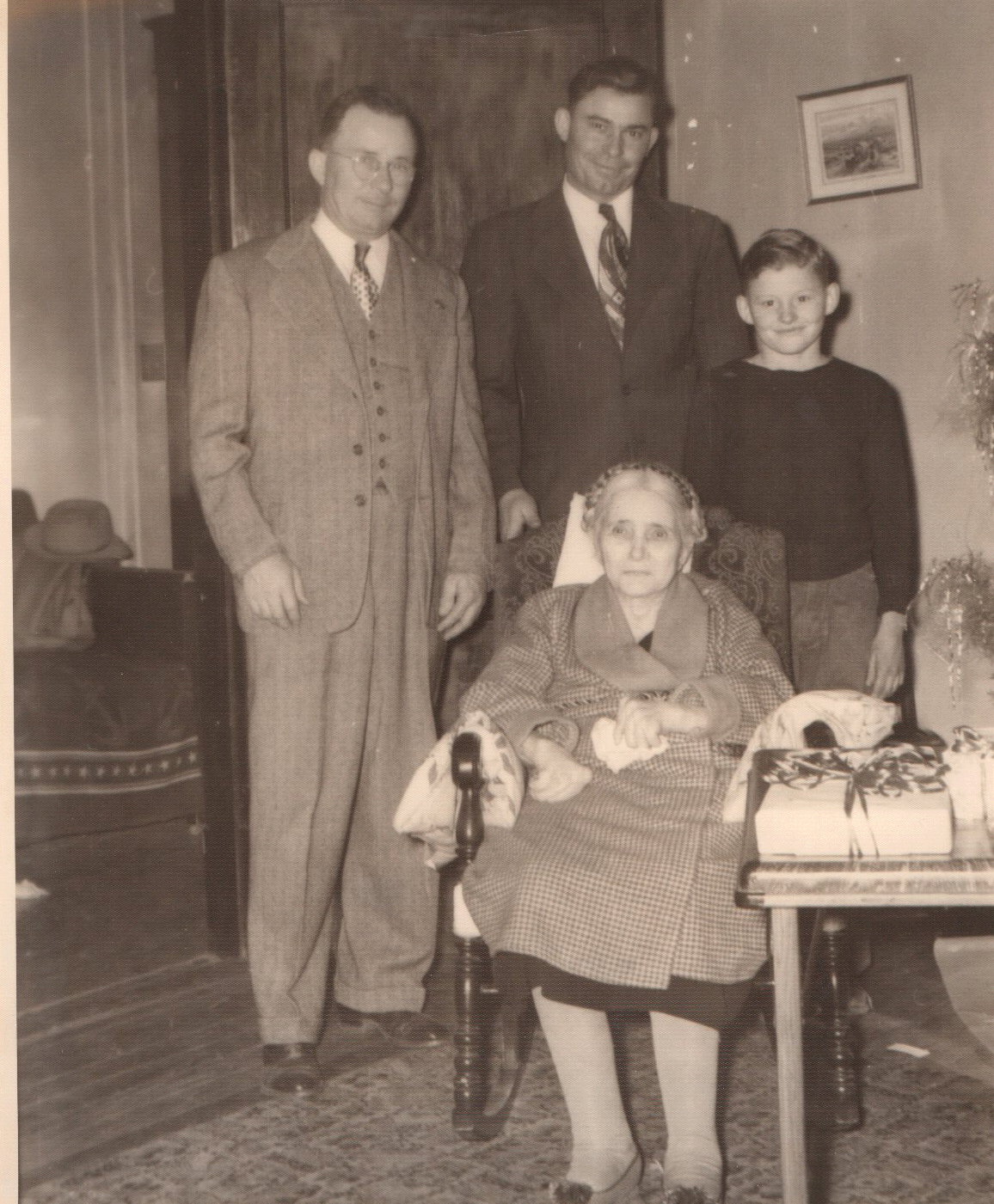 A black and white photo showing four men and a woman in a living room. The woman, elderly, is seated in a chair, and the men are standing behind her. The woman appears to be in a wheelchair, with a blanket on her lap. The room has a framed picture on