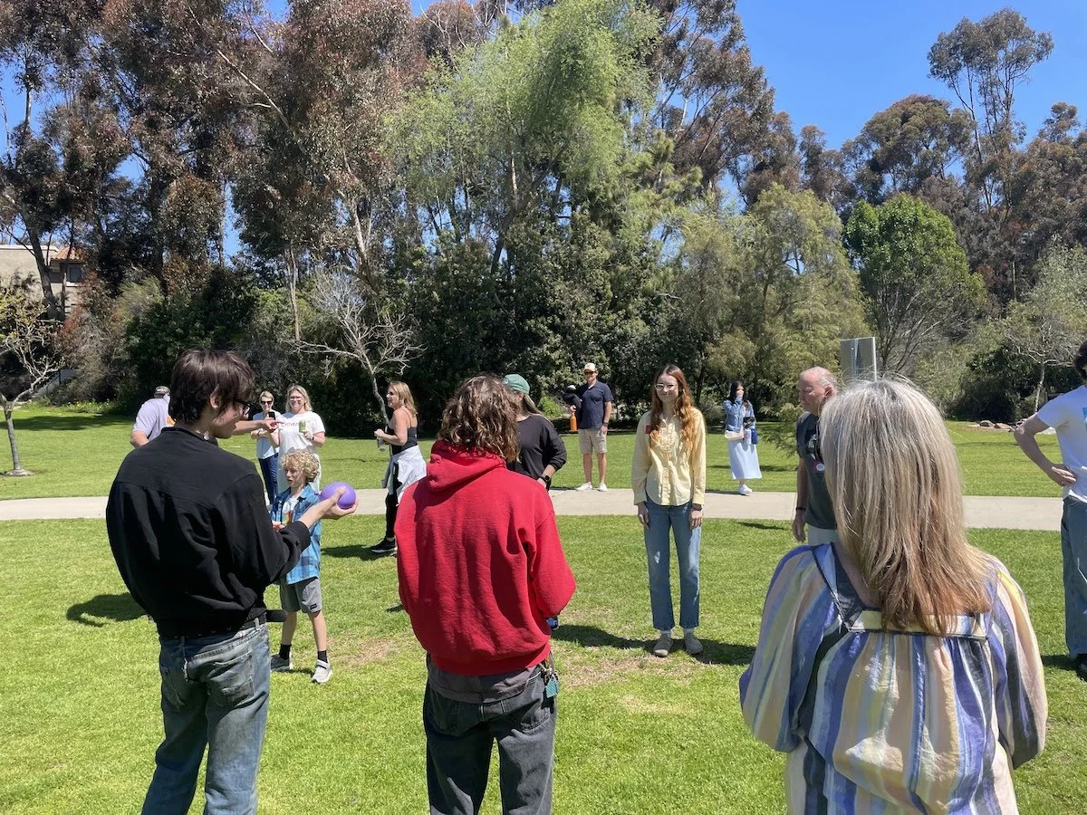 People gathered outdoors on a grassy park, some talking and one person holding a purple ball, with trees and a clear blue sky in the background.