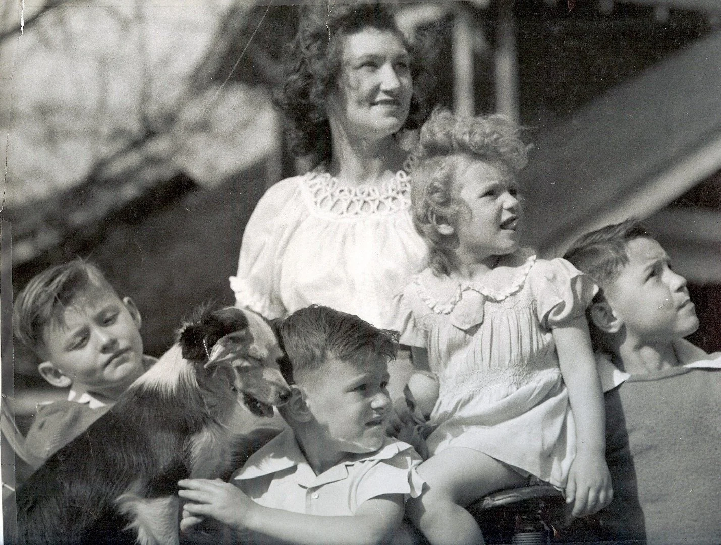 Black and white photograph of a woman with curly hair and light-colored dress sitting with four children and a dog, all looking in the same direction. The children appear to be sitting around her, and the scene seems to be outdoors.