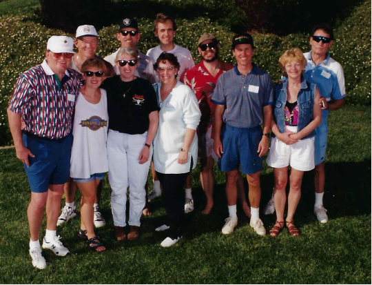 Group of ten people standing outdoors on grass, smiling, with trees and bushes in the background.