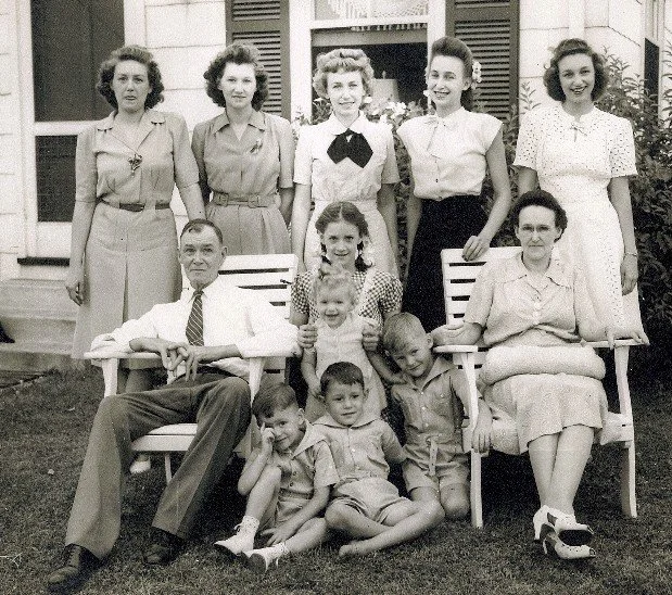 Group of nine people, including six women, one man, and four children, posing outdoors in front of a building with window shutters. Three people are seated on chairs, while the others stand behind them.