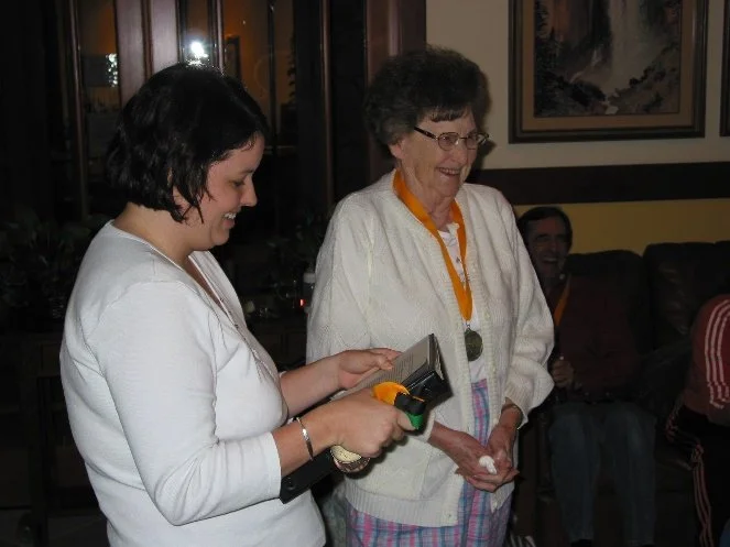A young woman and an elderly woman smiling and holding a certificate or paper in a cozy indoor setting, with other people sitting in the background.