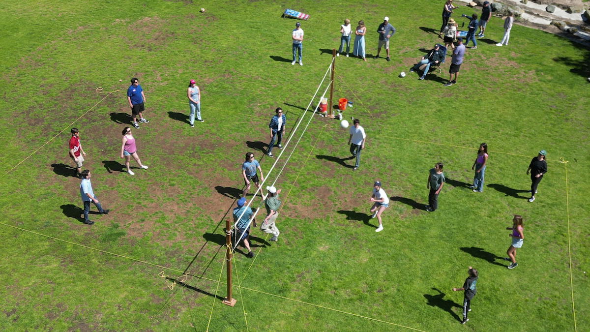 People playing volleyball on a grass field with some areas of patchy grass, surrounded by yellow boundary lines, and spectators watching from outside the court.