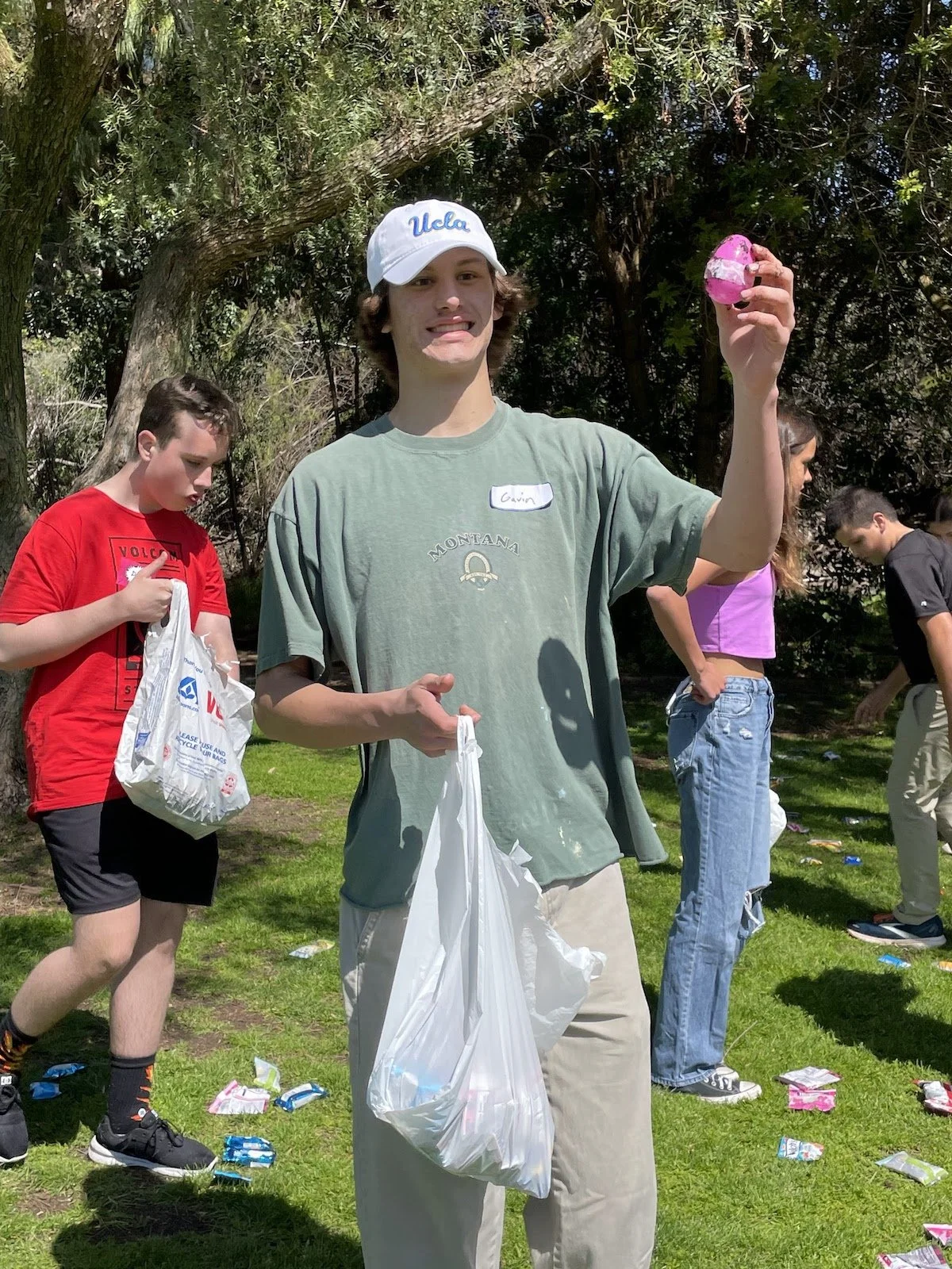 A young man wearing a UCLA cap and a green t-shirt holding a pink and black Easter egg, during a cleanup event outdoors with other participants and scattered candy wrappers on the grass.