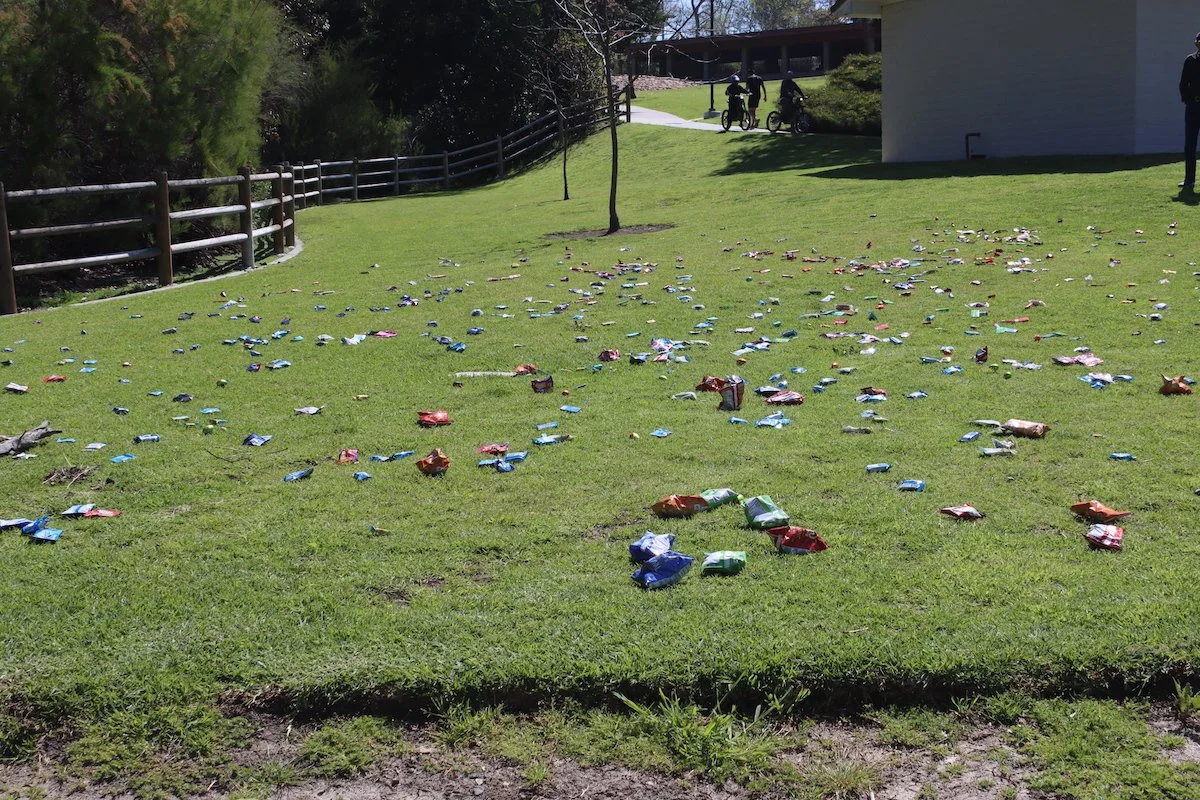 Litter of snack wrappers scattered on a grassy field with a fence, trees, a building, and people in the background.