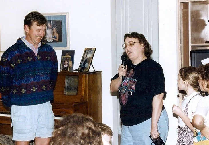 Woman speaking into a microphone while a man and children listen indoors.