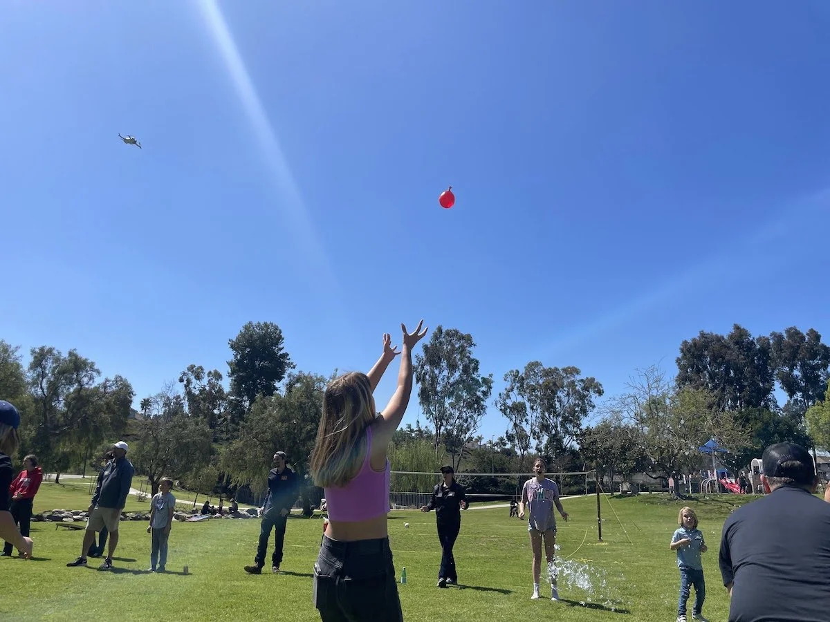 Group of people playing volleyball outdoors on a grassy field, with a blue sky and trees in the background. A girl in a pink top is reaching up to catch a red balloon, and a helicopter is flying in the sky.
