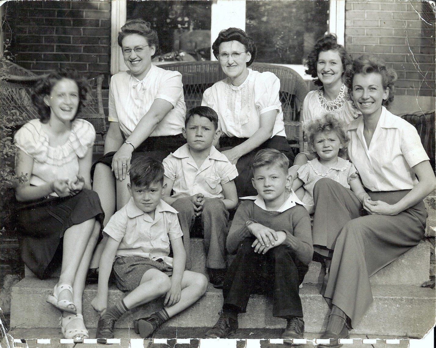 Black and white photograph of seven children and three women sitting and standing on steps outside in front of a brick building, smiling and looking at the camera.