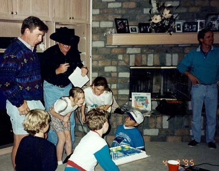 A group of children and adults gathered in a living room for a birthday celebration, with a cake and presents on the floor in front of them, and a brick fireplace and framed photos in the background.