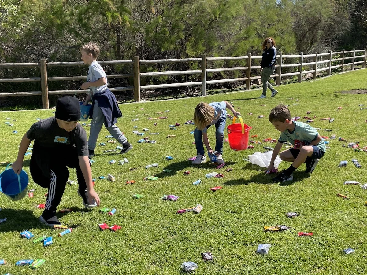 Children collecting candy on a grassy field with a woman nearby, fallen candy wrappers scattered all around, surrounded by trees and a wooden fence.