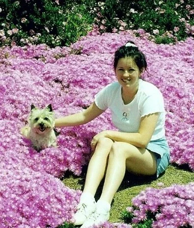 A woman sitting on pink azalea flowers in a garden with her small, white dog.