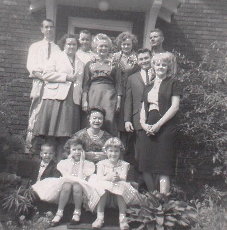 A black-and-white photo of a family group standing and sitting outside a house with a brick wall and plants surrounding them.